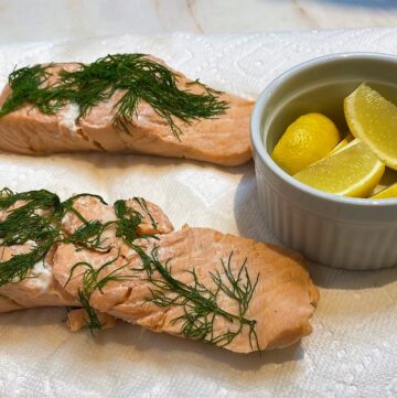 Cooked pink salmon filets topped with dill draining on white paper towels with a round white dish of yellow lemon wedges