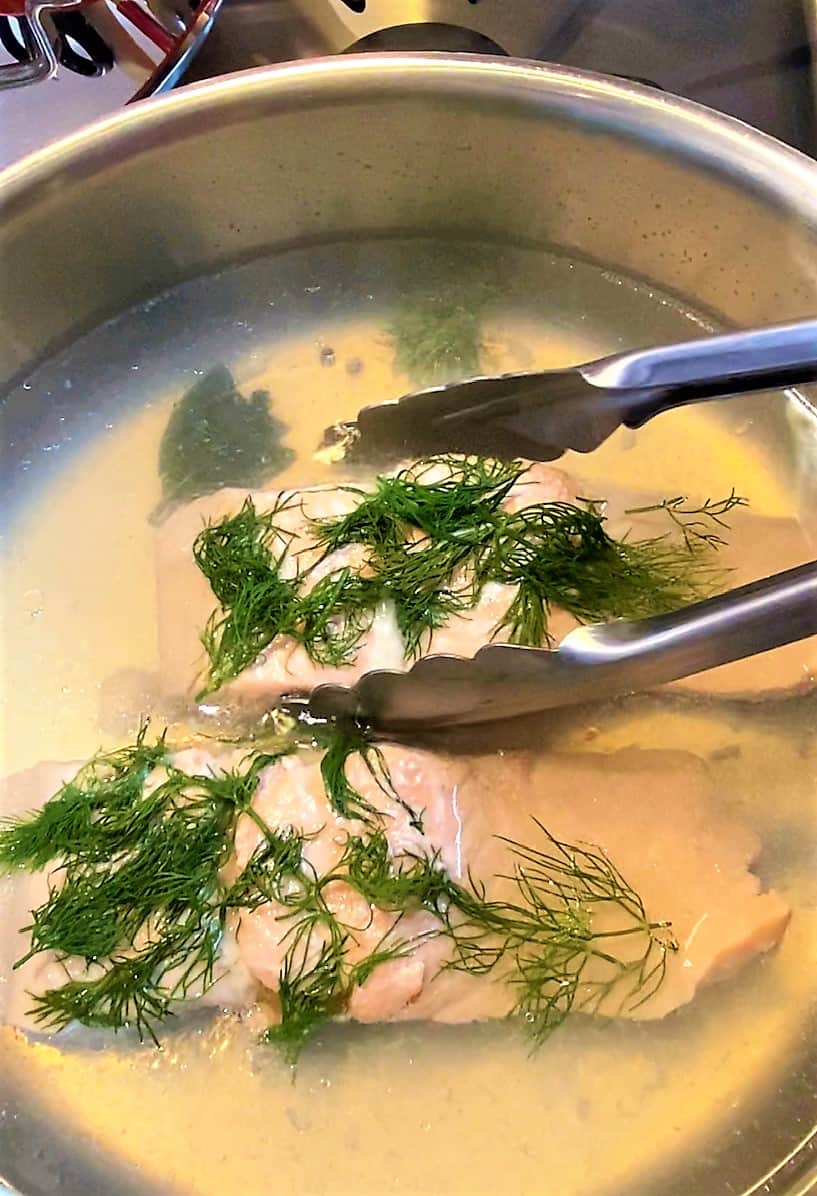 Tongs lifting pink salmon fillet with dill on top from poaching liquid in pan