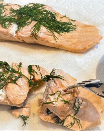 Cooked salmon fillets topped with dill on white paper towels being flaked with a fork