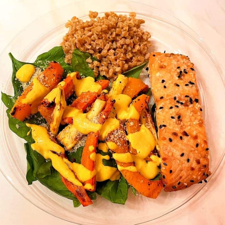 Cooked salmon fillet alongside spinach topped with root veggies and yellow turmeric dressing and scoop of farro.