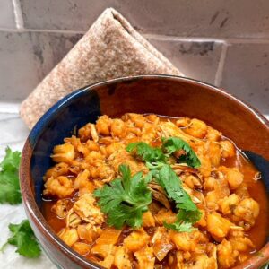 A folded wheat tortilla sits behind a rustic bowl full of orange-red Pork Pozole stew garnished with a green cilantro spring.