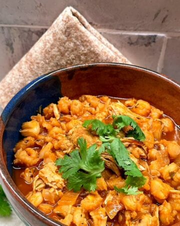 A folded wheat tortilla sits behind a rustic bowl full of orange-red Pork Pozole stew garnished with a green cilantro spring.