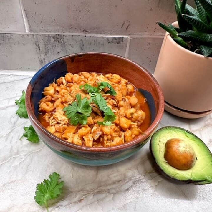 An orange-red bowl of pork pozole featuring hominy corn garnished with cilantro and half an avocado off to the side.