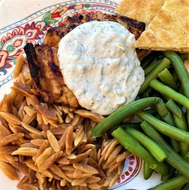 savory Greek yogurt sauce topping grilled chicken thigh with wheat orzo, green beans, and pitas on paisley plate