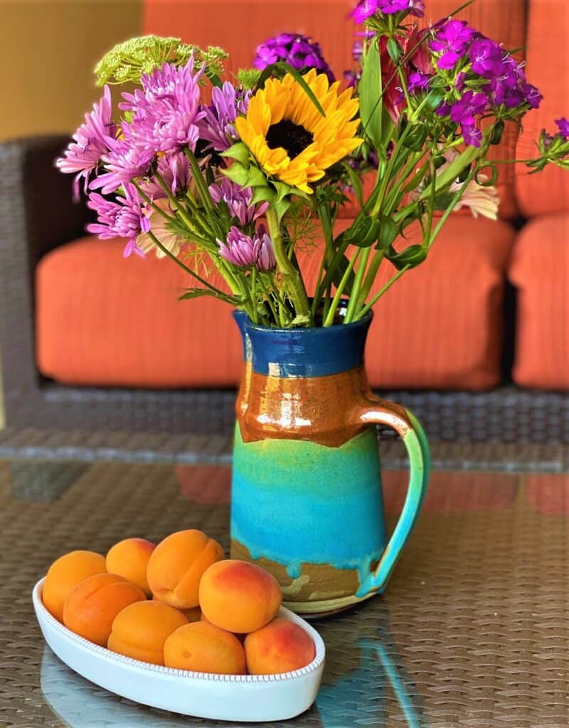 A dish of ripe apricots with a colorful pitcher of summer flowers