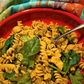 Creamy, curried, chickpea rotini with spinach and mushrooms in a red dish with a colorful kitchen towel