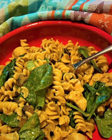 Creamy, curried, chickpea rotini with spinach and mushrooms in a red dish with a colorful kitchen towel