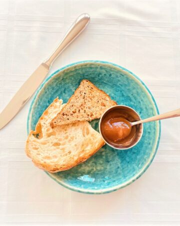 Silver ramekin with apple butter and petite spoon with toasted bread on blue-green plate, white cloth background with knife