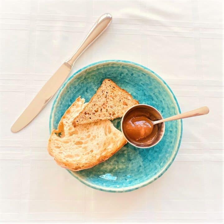 Silver ramekin with apple butter and petite spoon with toasted bread on blue-green plate, white cloth background with knife