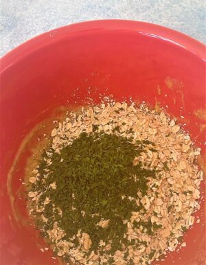 Dried parsley, whole oats, and oat flour added to wet ingredients for dog treats in red bowl