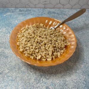 Orange bowl with scalloped edges filled with cooked barley on blue background; serving spoon inserted in the barley