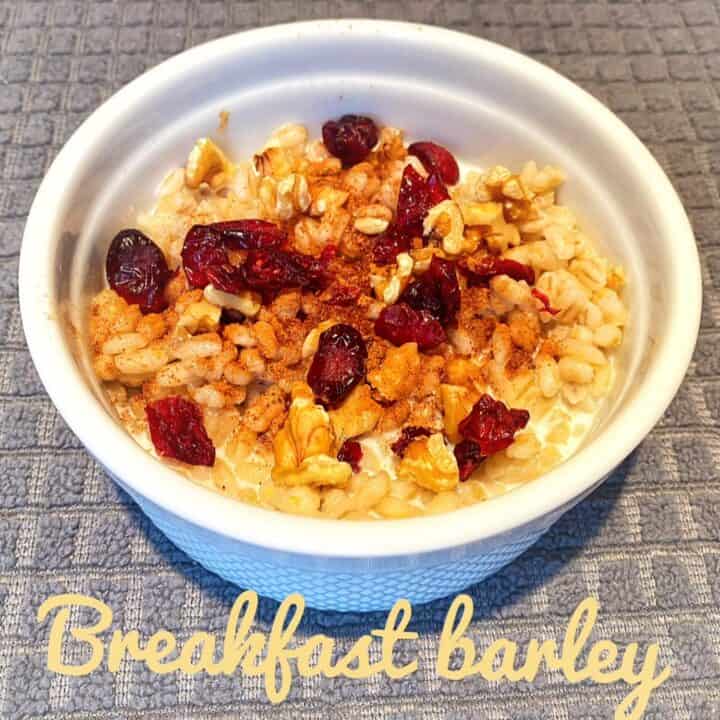 White bowl of breakfast barley topped with craisins, milk, nuts, and spices on a gray background