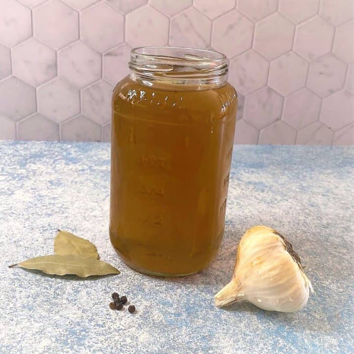 quart jar full of veggie scraps broth with bay leaves, peppercorns, and a head of garlic on a blue countertop with pink hex backsplash