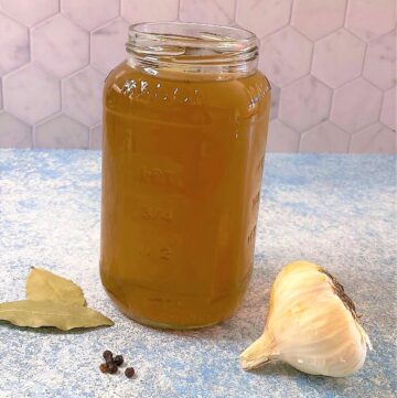 Mason jar with veggie scraps broth on a blue countertop with garlic, black peppercorns, and two bay leaves