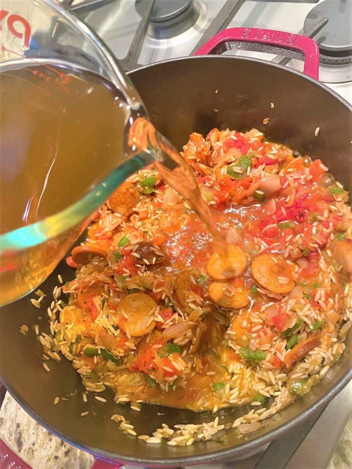 Pouring broth into the healthy jambalaya cooking in Dutch oven over a gas cooktop