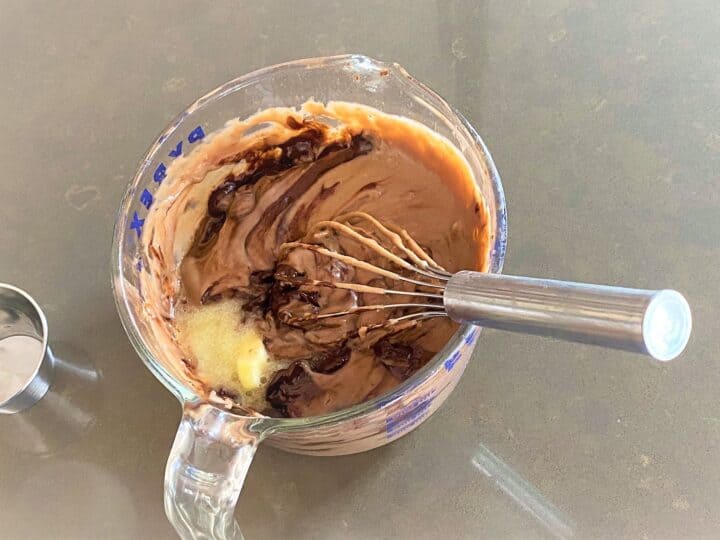 Whisking melty chocolate chips, butter, and almond extract into hot chocolate pudding in a large glass measuring cup on brownish background