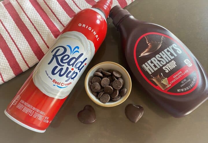 reddi-whip can, hershey's chocolate syrup, chocolate chips, and chocolate hearts on brown background with red and white striped towel