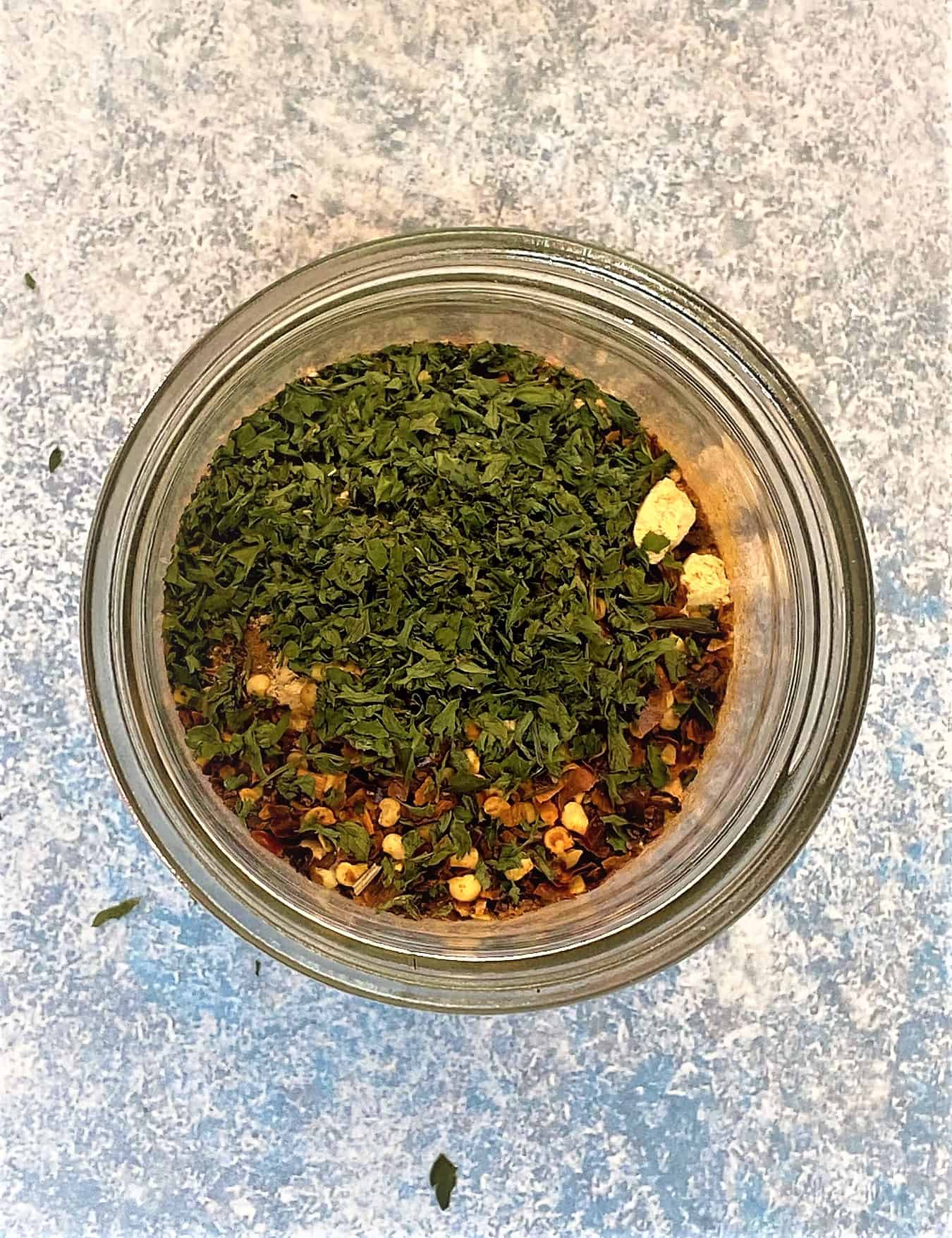 round jar on blue mottled background with spices and a top layer of greed dried parsley