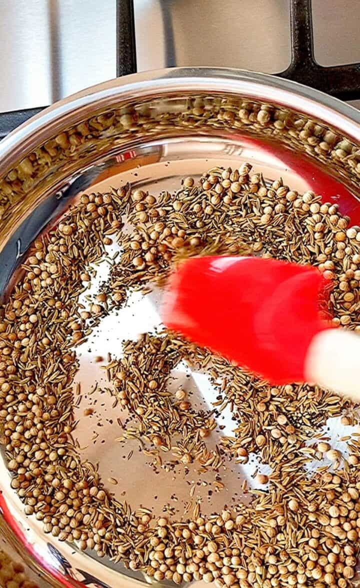 Black pepper, coriander and cumin seeds in shiny skillet with red spatula pushing them around