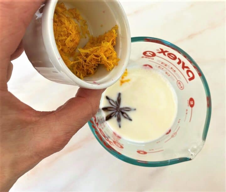 Star anise pod floats in milk in a measuring cup while orange zest is begin added