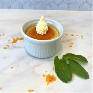 Orange pudding cake in white ramekin on white marble countertop with orange zest and mint leaves around it