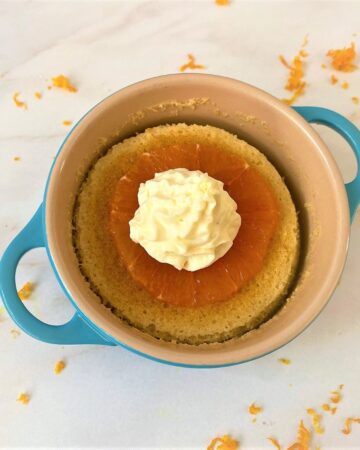 Orange pudding cake with star anise in a teal ramekin topped with cross section of orange and piped whipped cream on white marble countertop