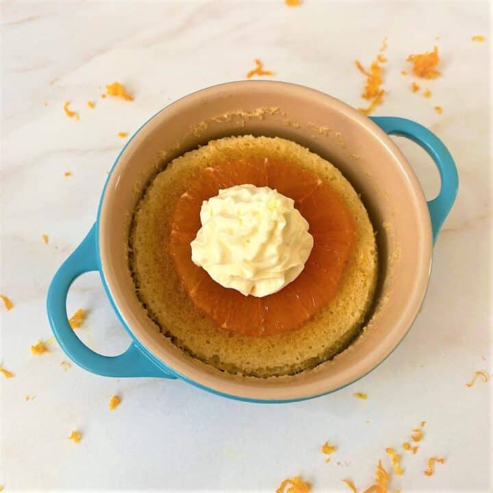 Orange pudding cake with star anise in a teal ramekin topped with cross section of orange and piped whipped cream on white marble countertop