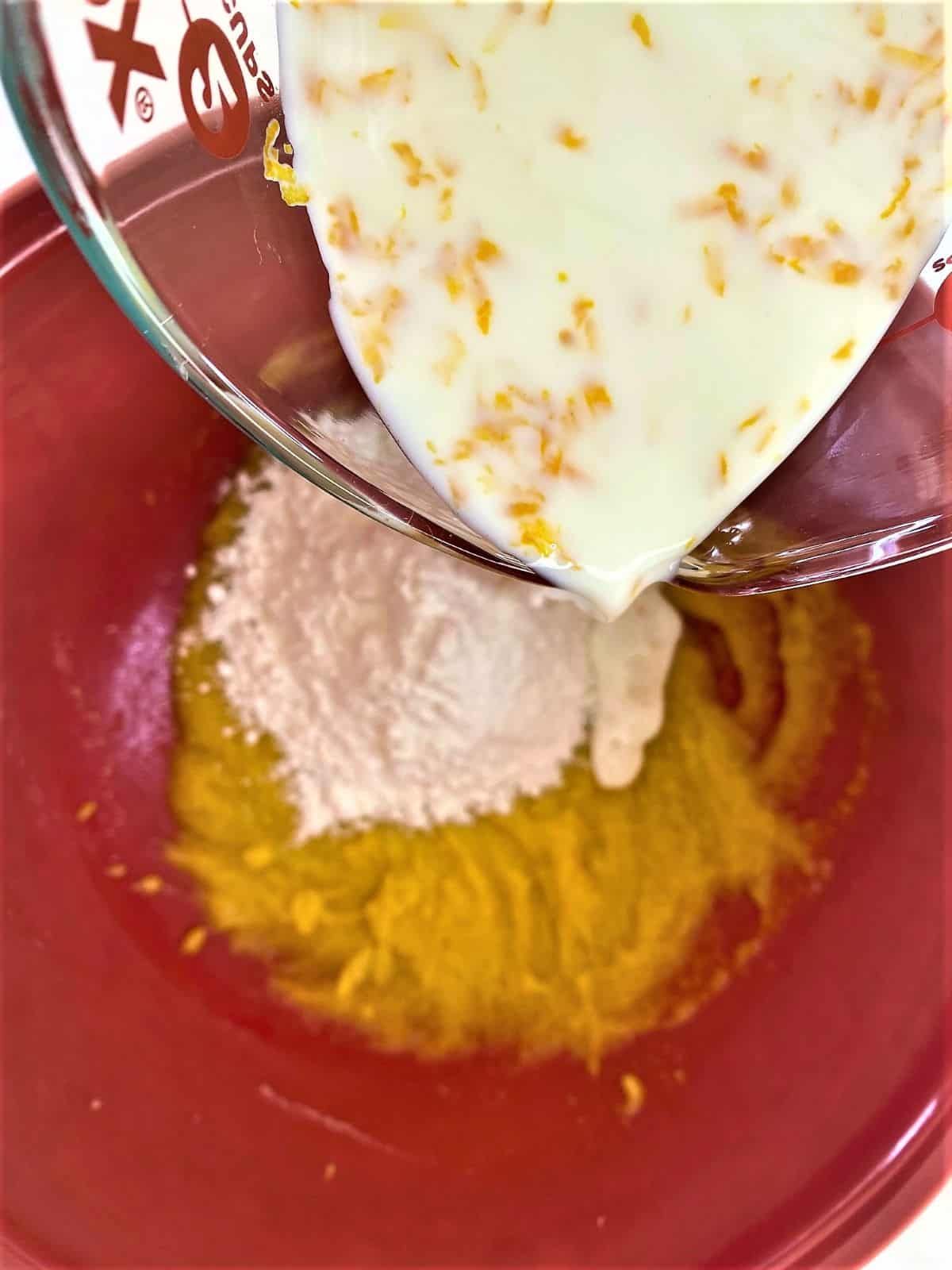 adding milk with orange zest in it to red mixing bowl that holds flour and yolk mixture