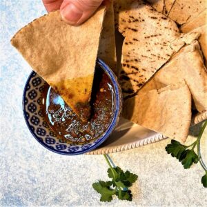 Pita wedge emerging from Chermoula Dip with red-brown oil-based dip covering tip of pita on a mottled blue backgroundtip