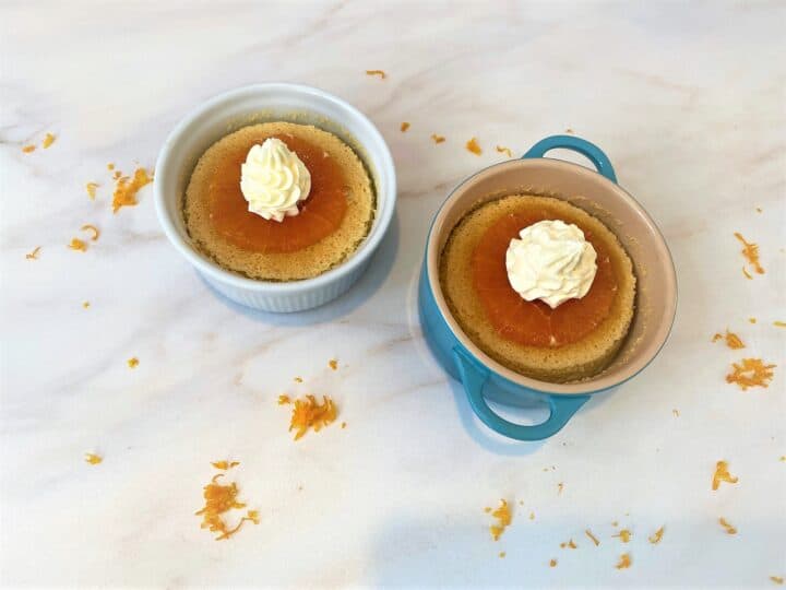 individual orange pudding cakes in white and teal ramekins garnished with orange cross section and whipped cream on marble countertop