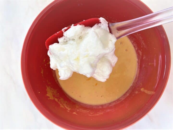 Spatula with beaten whites going into a red bowl with yellow batter