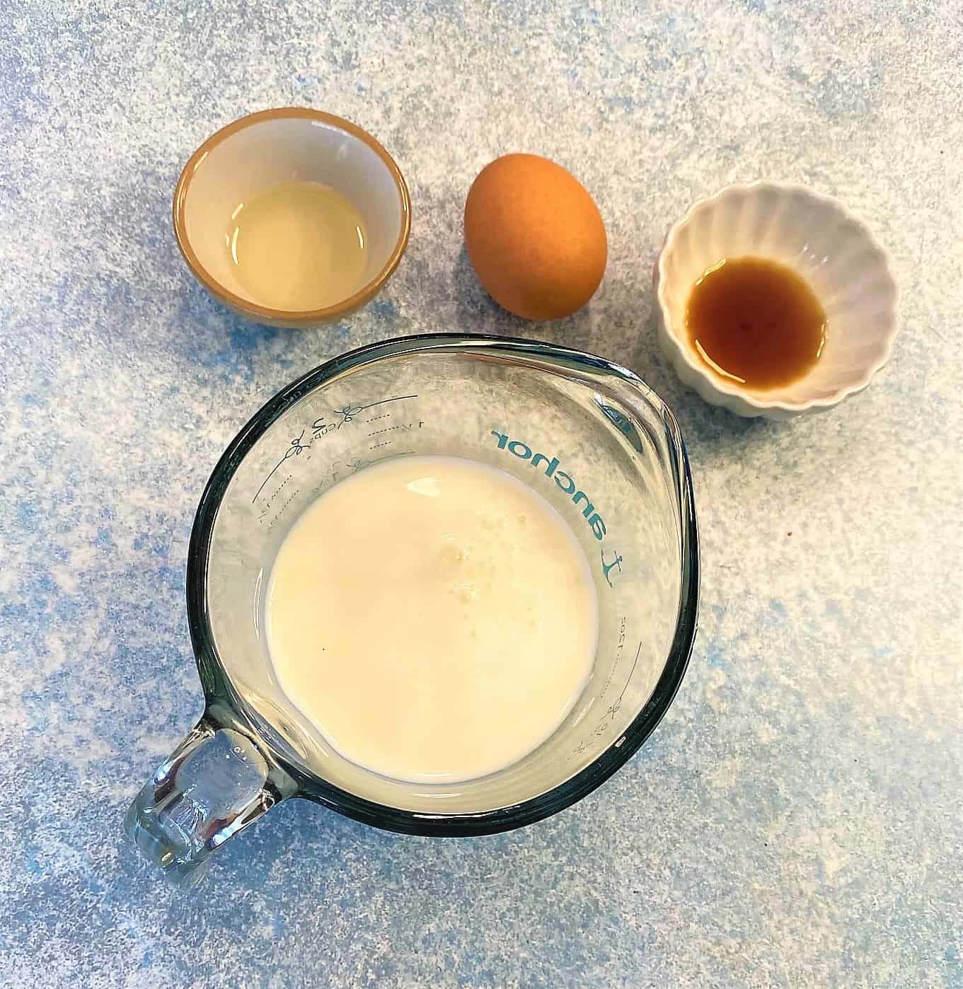 Cup of milk in glass measuring cup with vanilla, brown egg, and vegetable oil on mottled blue countertop