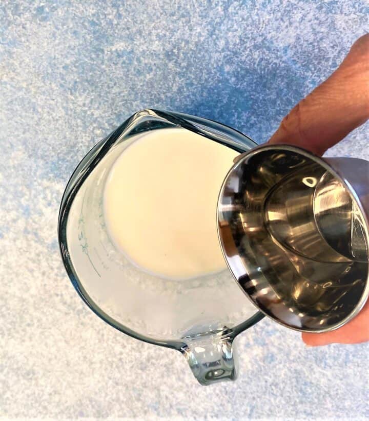 Small amount of clear vinegar being poured into a milk in a measuring cup on a bluish mottled countertop