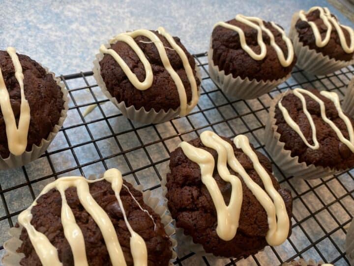 chocolate muffins topped with white chocolate drizzle on a baking rack on a blue countertop