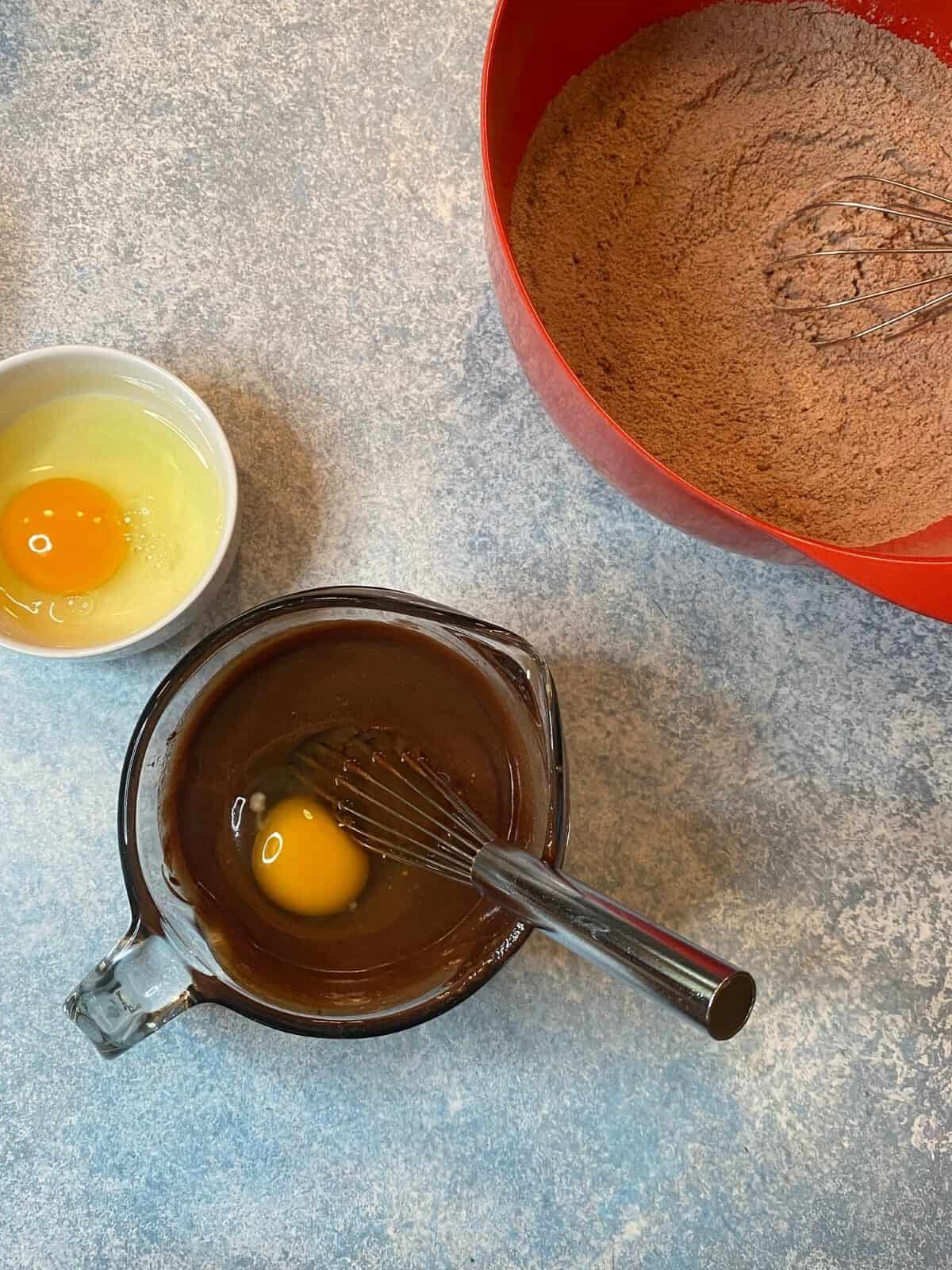 Raw egg in white bowl and raw egg in melty chocolate in measuring cup on blue countertop