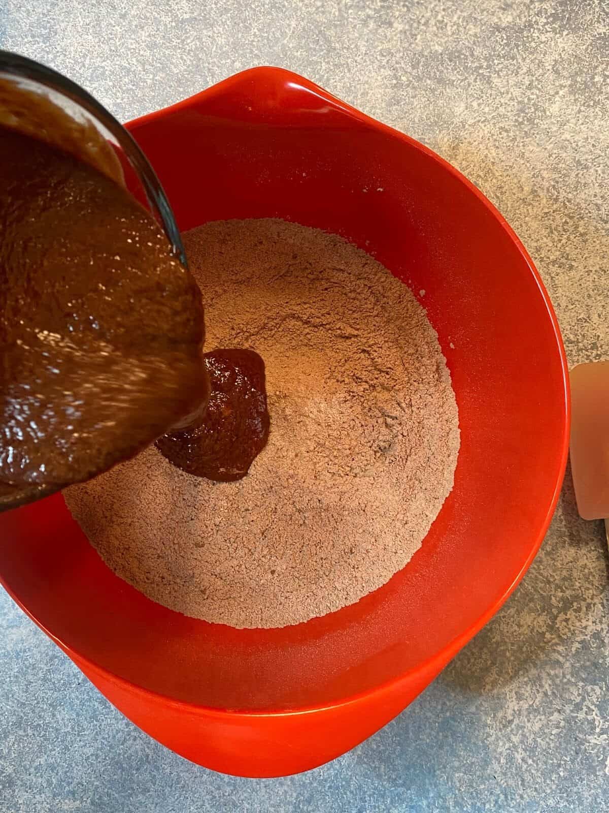 Pouring a chocolatey looking mixture into dry chocolatey ingredients in a red bowl