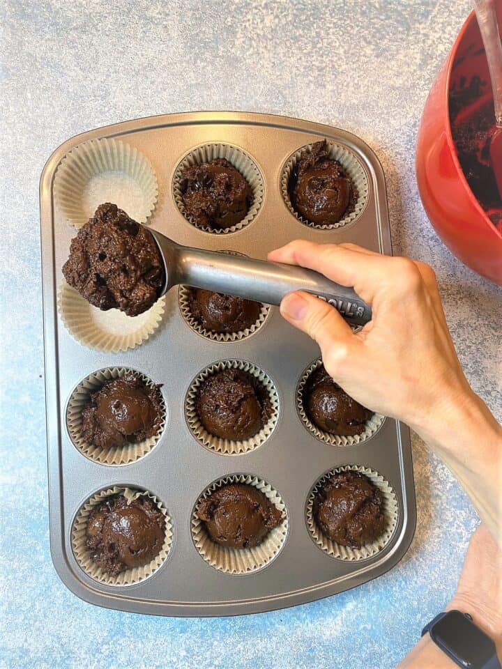 Muffin tin with chocolatey batter being scooped into muffin liners with an ice cream scoop