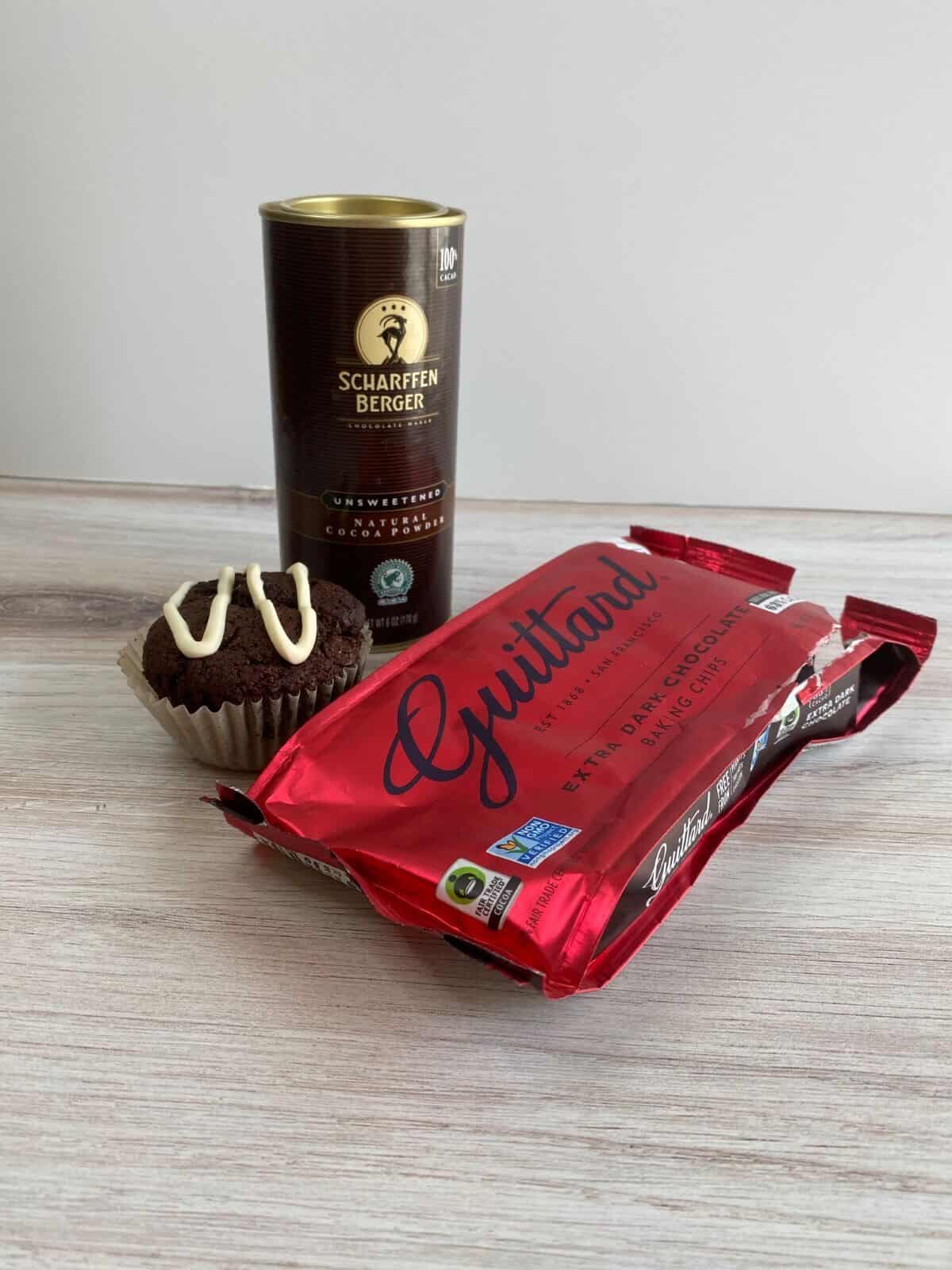 Red Guittard dark baking chip wrapper, chocolate muffin with white drizzle, Scharffen Berger cocoa tin on light wood grain surface