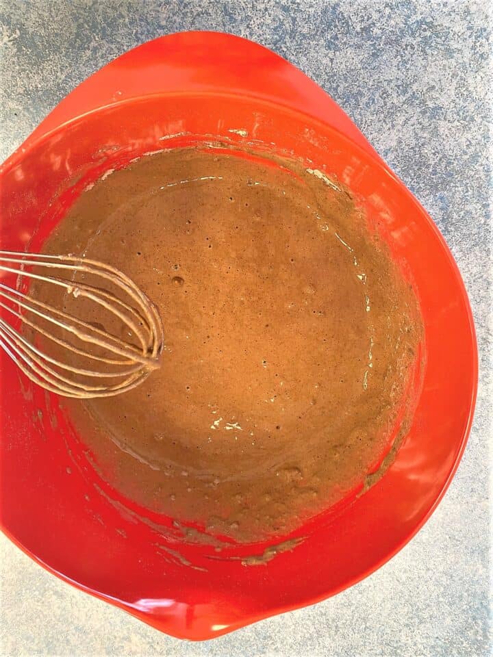 Chocolatey batter for protein pancakes in red bowl with whisk, mottled bluish countertop