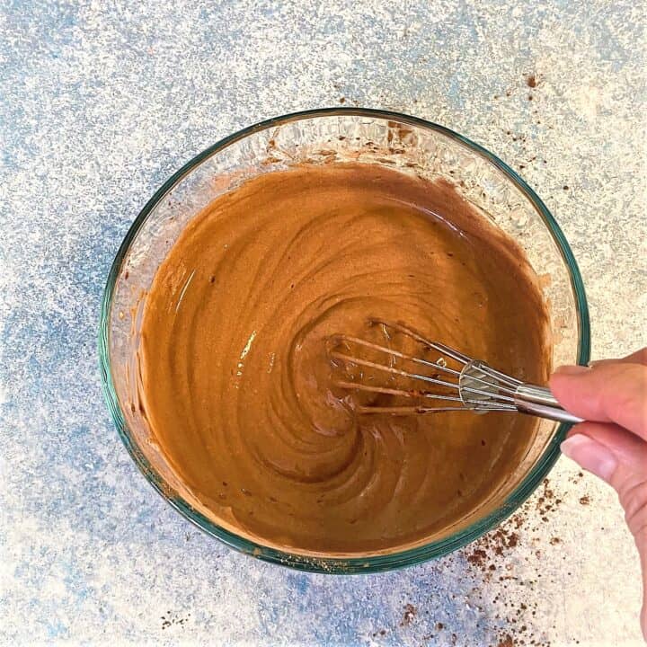 Thick chocolate yogurt sauce in clear bowl being whisked on blue mottled countertop