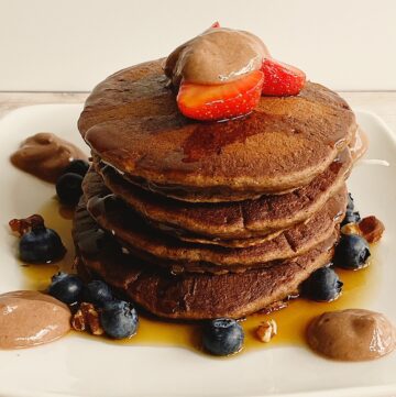 stack of fluffy chocolate protein pancakes with blueberries and strawberry slices, chocolate yogurt sauce and syrup