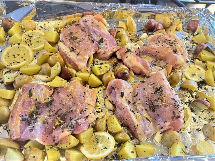 Raw chicken thighs on sheet pan with potato chunks and lemon and seasonings