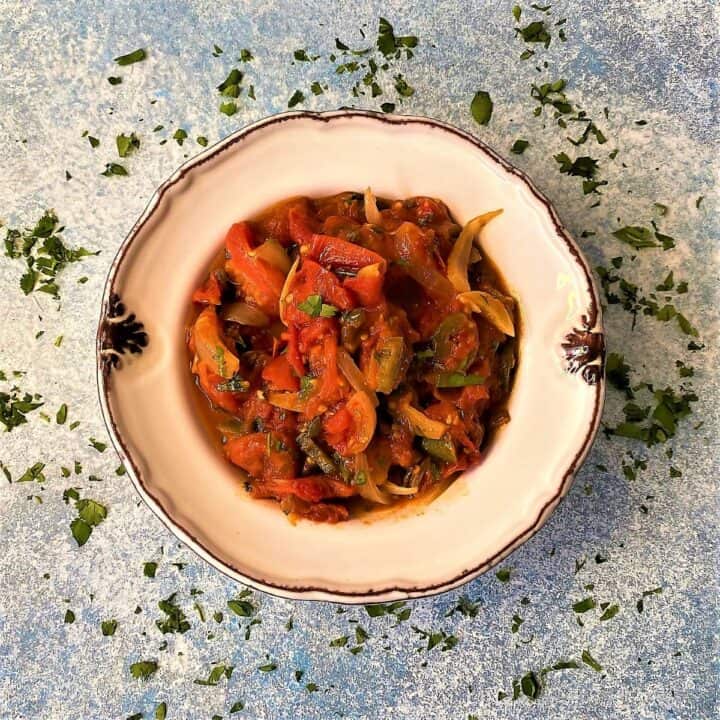 Pretty white ceramic bowl filled with chunky Roasted Tomato Chutney with chopped herbs sprinkled around it on a mottled blue background