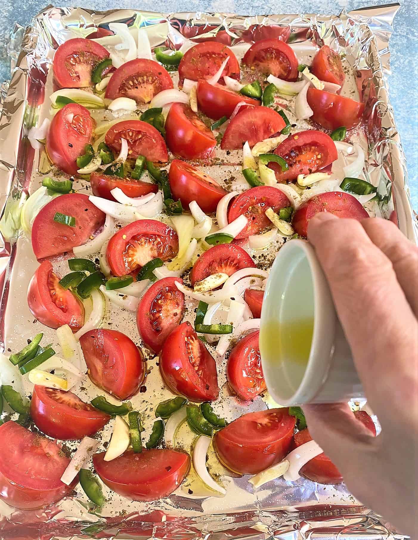 Pouring olive oil from a small white cup onto tomato wedges, green pepper, onion, garlic on foil-lined sheet pan