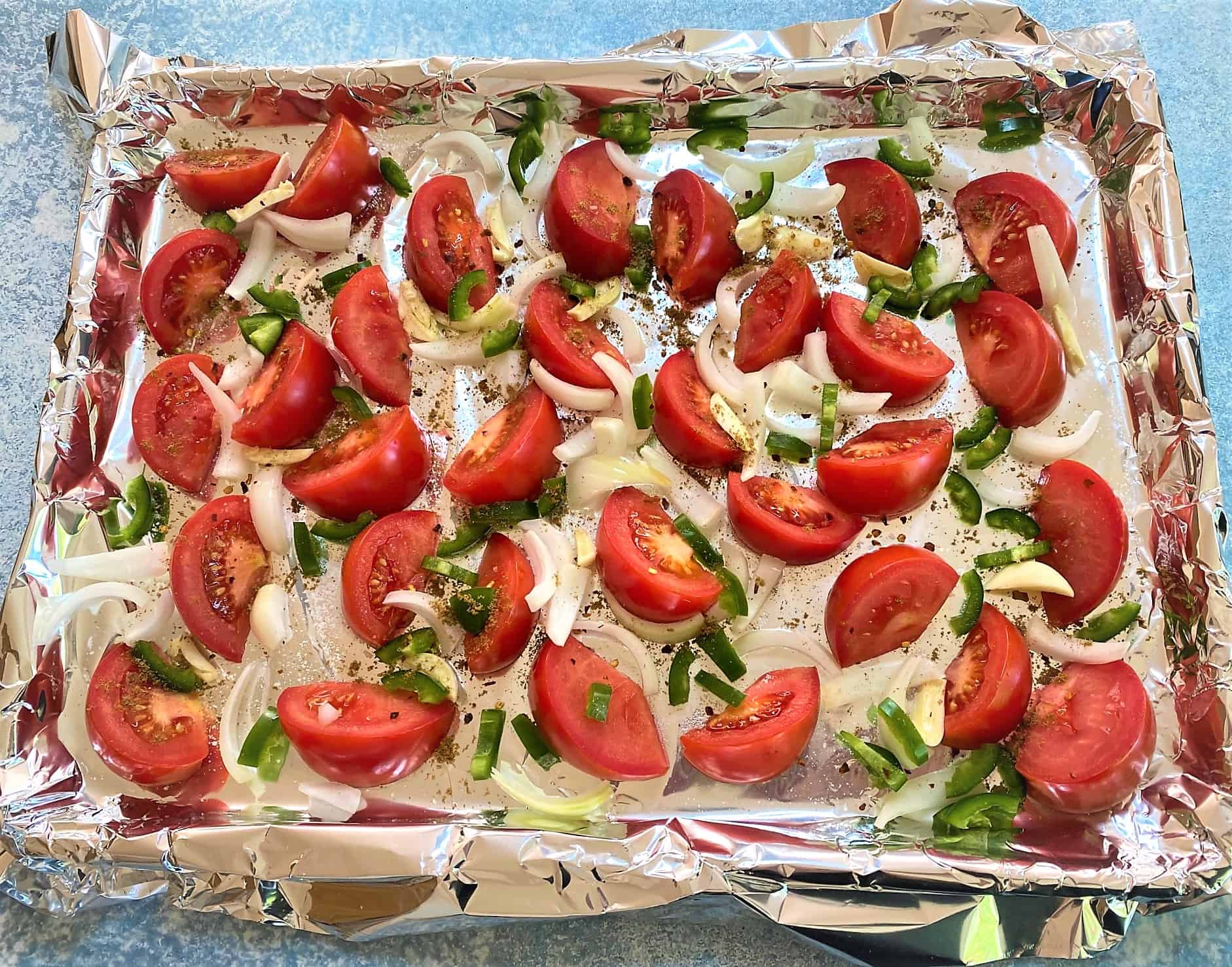 Tomato wedges, onion and jalapeno bits, along with garlic pieces and spices on a foil-lined sheet pan