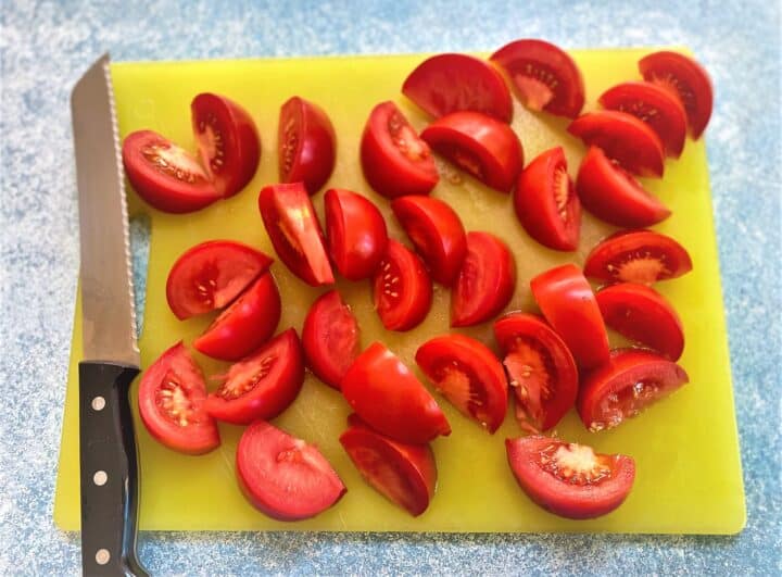 Yellow cutting board covered with tomato wedges with serrated knife nearby on mottled blue background