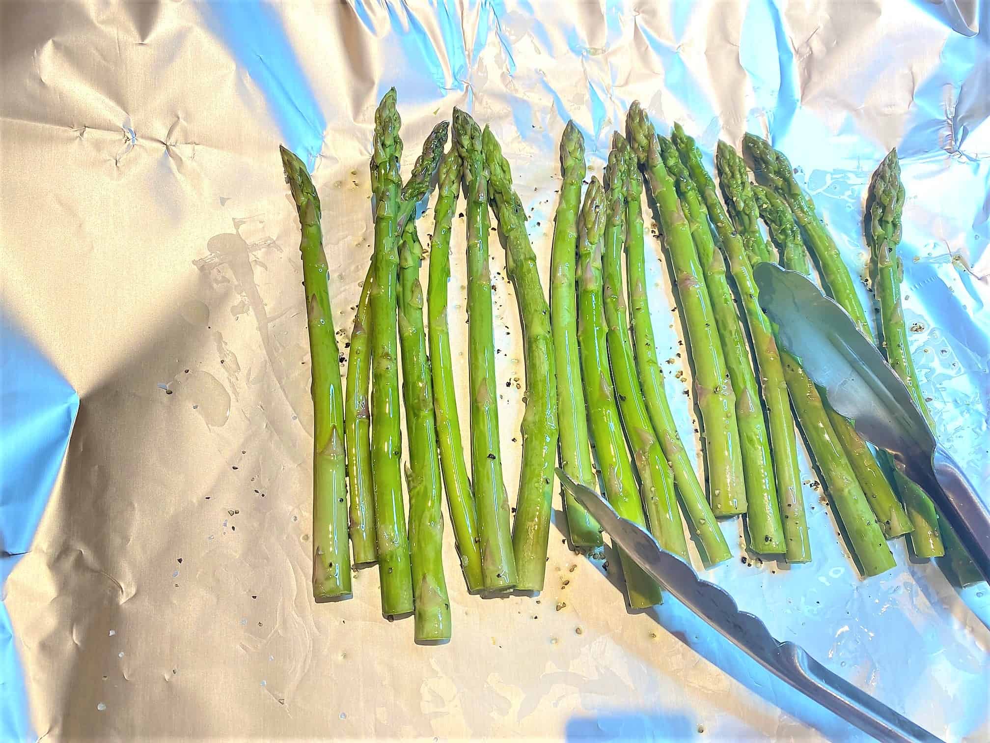 Tongs tossing asparagus in seasonings on shiny foil