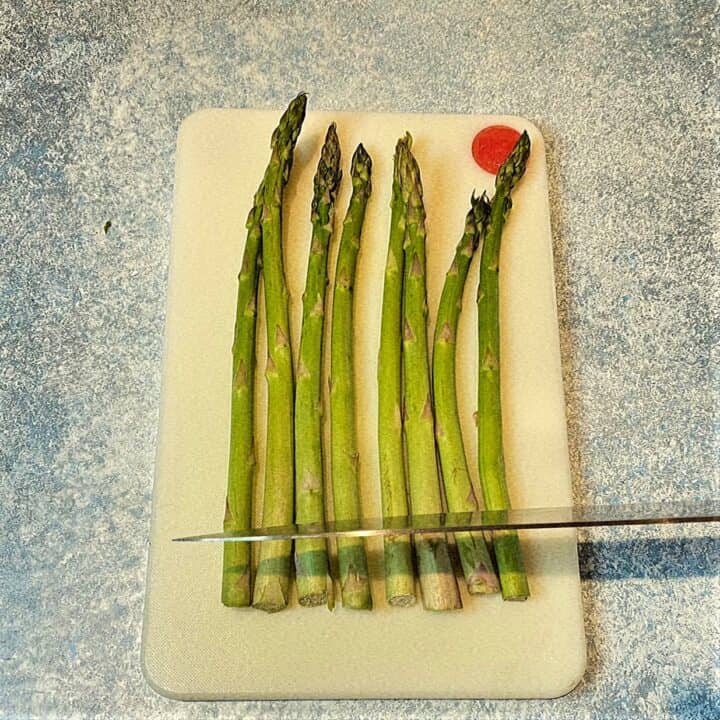 Asparagus lined up on a white cutting board with chef's knife cutting off the bottoms; on a mottled blue countertop