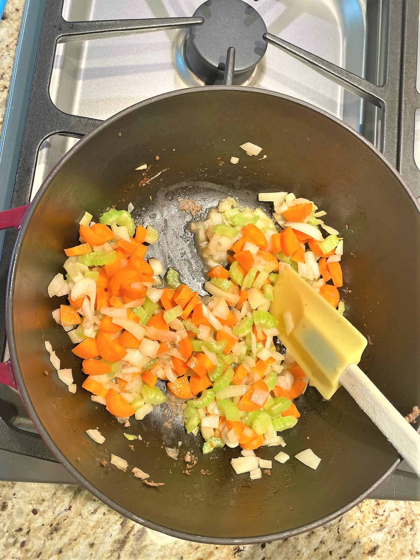 Wood and green silicone spatula stirring Chopped onion, carrot, and celery cooking in dark Dutch oven on stovetop