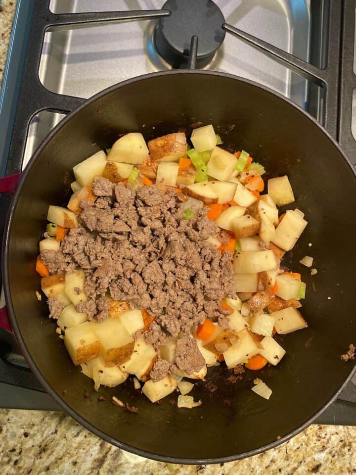 Crumbled turkey sausage atop briefly cooked potato cubes and chopped onion, celery, and carrot in dark Dutch oven on stovetop in tan and beige granite countertop